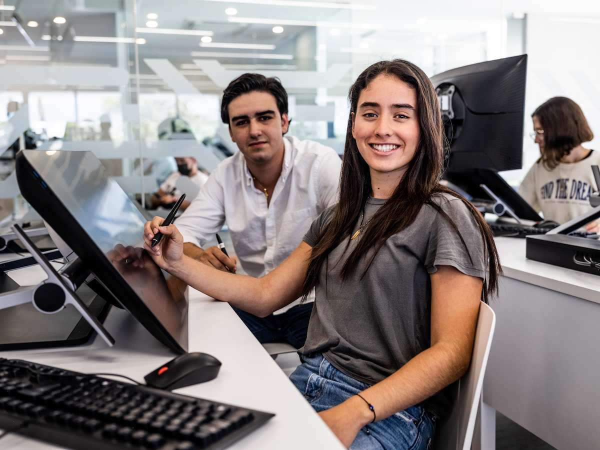 Estudiantes trabajando con lápices ópticos en pantallas gráficas en un aula tecnológica con fondo de cristal.